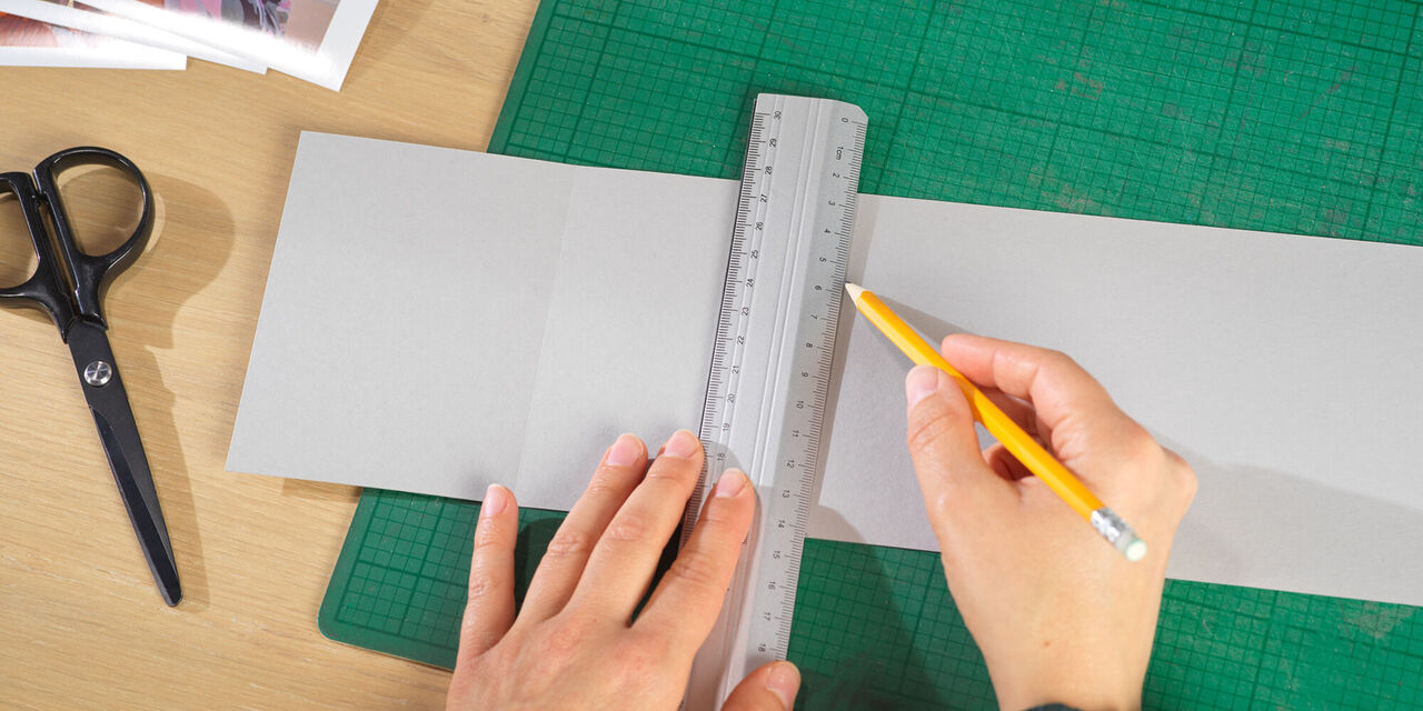 Két kéz ceruzával és vonalzóval vonalakat húz a fotók számára egy szürke kartonlapra.