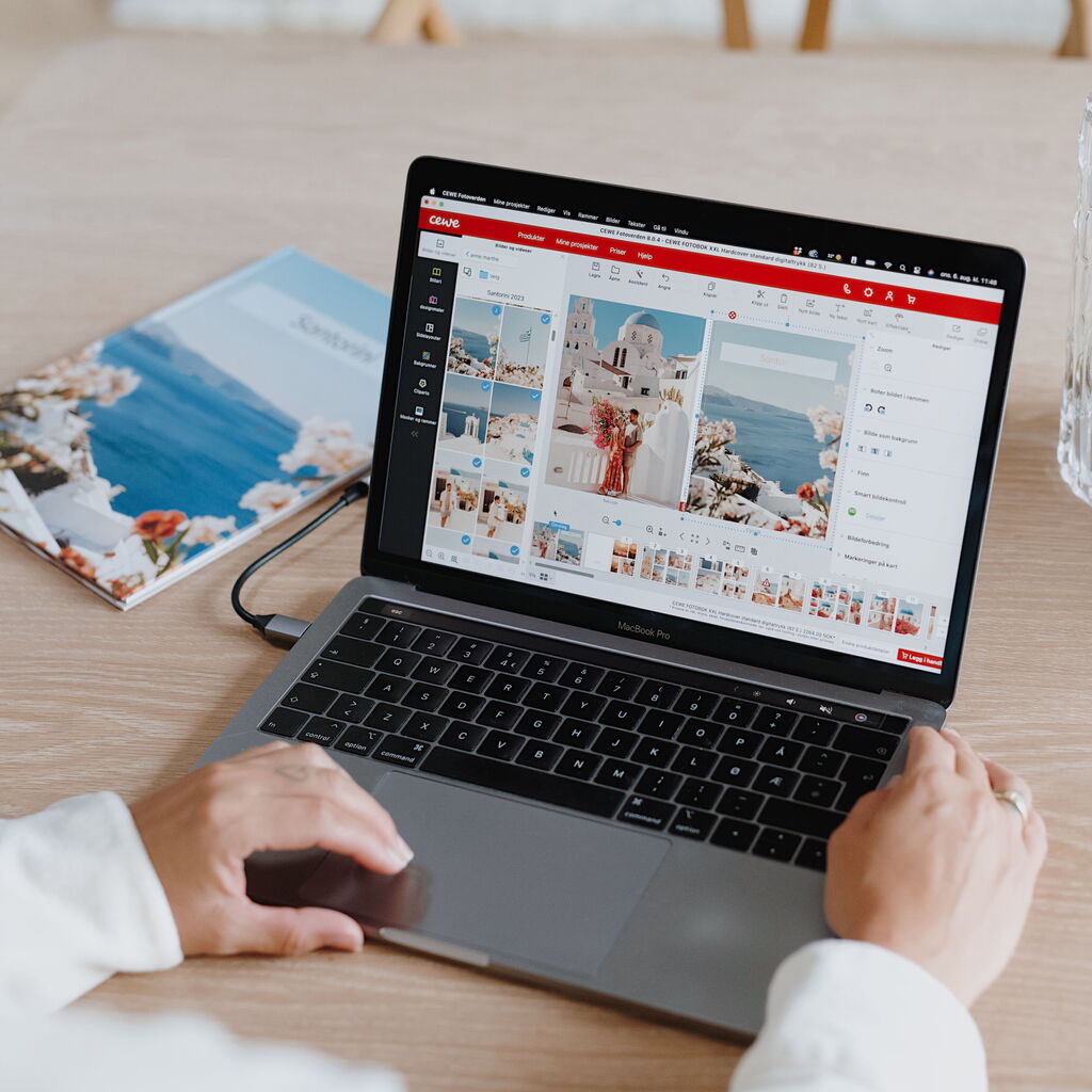 Egy ember egy asztalnál ülve laptopon dolgozik. A képernyőn a CEWE Fotóvilág szoftver látható, benne egy nyaralás képeivel.
