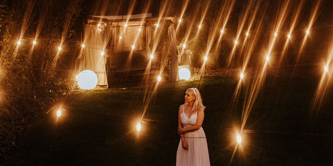 Egy fehér ruhás nő áll az éjjeli szabad ég alatt. Hangulatvilágítás veszi körül, a háttérben egy kivilágított pavilon áll.